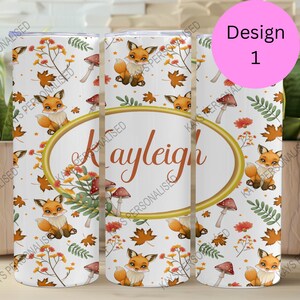 May include: A white tumbler with a design of orange foxes, mushrooms, and autumn leaves. The name "Kayleigh" is in a gold oval. The design is labelled "Design 1" in a pink circle.