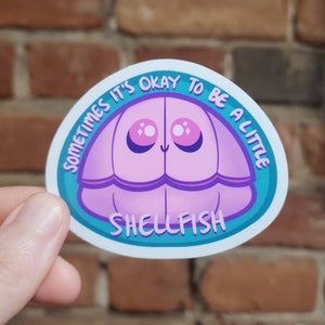 May include: A teal blue and white sticker with a pink cartoon clam with sparkly eyes. The text on the sticker reads "Sometimes it's okay to be a little shellfish".