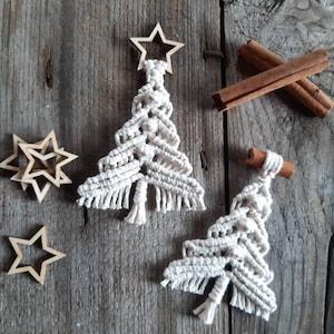 Può includere: Due decorazioni per l'albero di Natale in macramè bianco con stelle in legno in cima su uno sfondo in legno rustico. Le decorazioni sono fatte a mano e presentano un dettaglio di frangia in basso.