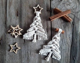 Puede incluir: Dos adornos de árbol de Navidad de macramé blanco con estrellas de madera en la parte superior sobre un fondo de madera rústico. Los adornos están hechos a mano y tienen un detalle de flecos en la parte inferior.