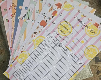 May include: A stack of punched paper inserts for a binder, featuring various colourful patterns including lemons, autumn leaves, and floral designs. The top sheet is a Sinking Funds tracker with German text headings and a monthly savings table.