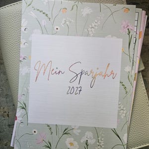 Könnte beinhalten: Ein salbeigrüner Einband für einen Planer mit einem zarten Blumenmuster aus weißen, rosa und gelben Wildblumen. In der Mitte steht der Text Mein Sparjahr 2027 in Schreibschrift, unten ist der Markenname FallinLoveDE aufgedruckt.