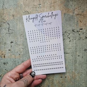 Könnte beinhalten: Eine weiße Karte mit dem Text "Kleingeld Sparchallenge" und Zahlenwerten zum Sparen. Die Karte wird von einer Hand mit einem Totenkopf-Design auf einem Fingernagel gehalten. Der Hintergrund ist eine strukturierte, gedämpfte grüne Oberfläche.