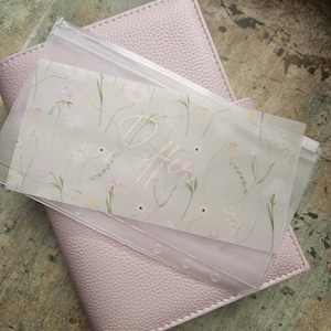 Könnte beinhalten: Ein hellvioletter Planer mit strukturierter Hülle, mit einer transparenten Kunststofftasche mit floralem Design und dem Wort "Buffer". Die Tasche enthält Bargeldumschläge, ideal für die Budgetierung und Organisation der Finanzen.