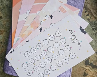 May include: A stack of rectangular paper cards featuring a 26-week challenge tracker with numbered circles from 1 to 26, decorated with small colourful hearts, resting on a textured lavender purple leather-like notebook cover.