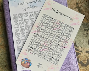 May include: Two rectangular paper savings challenge cards with the text Geldwäsche and Sparchallenge, featuring black line art of washing machines and laundry baskets, layered on top of a textured light purple leather-like binder cover.