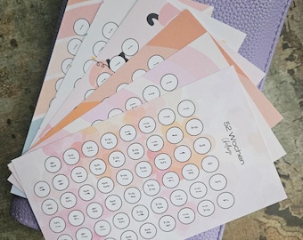 May include: A stack of rectangular paper challenge trackers with pastel watercolour backgrounds and numbered circles from 1 to 52, resting on a textured lavender leather-like binder. The top sheet features the text 52 Wochen Challenge.