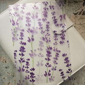 Può includere: Un inserto per planner trasparente con un motivo floreale di lavanda viola e verde. L'inserto è posizionato su un planner bianco strutturato con chiusura a bottone. Lo sfondo è una superficie in legno.