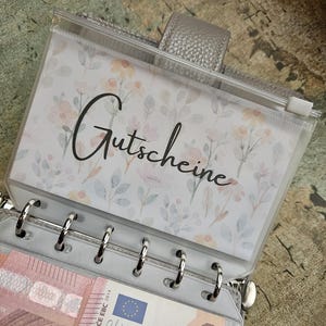 Könnte beinhalten: Ein grauer Ringbuchordner mit sechs Ringen und einer transparenten Reißverschlusstasche mit Blumenmuster, auf der das Wort Gutscheine in schwarzer Schreibschrift steht. Ein Zehn-Euro-Schein ist in einer Klarsichthülle sichtbar.