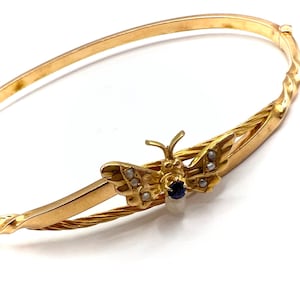 Könnte beinhalten: Ein goldenes Armreif-Armband mit einem Schmetterlingsanhänger. Der Schmetterling hat einen blauen Saphir in der Mitte und ist mit kleinen weißen Perlen verziert.