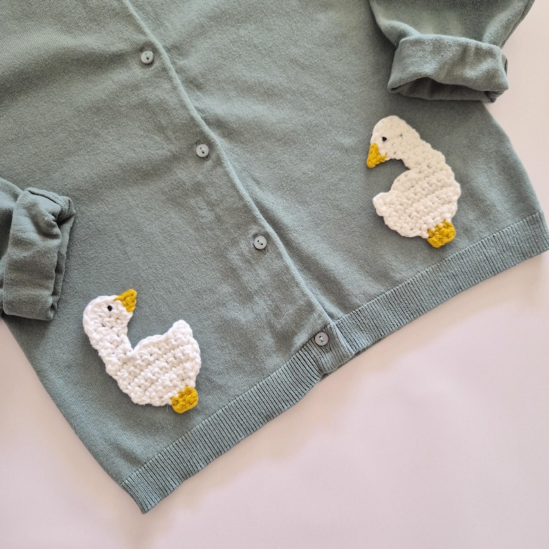 May include: A sage green knit cardigan featuring a button-down front and long sleeves with folded cuffs. Two white crocheted goose appliqu&eacute;s with yellow beaks and feet are attached to the lower front panels of the garment.