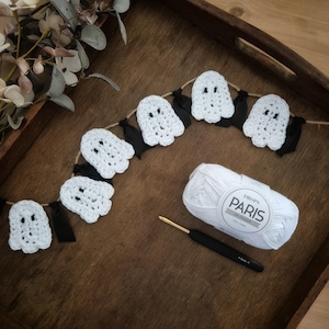 May include: A handmade Halloween garland featuring crocheted white ghost figures with black ribbon accents. A ball of white yarn, a crochet hook, and decorative foliage are also present, suggesting a craft project.