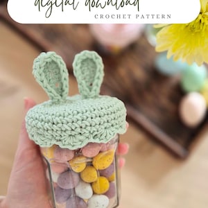 May include: A clear glass jar filled with colorful speckled candies, topped with a light green crocheted bunny ear lid. The text "digital download CROCHET PATTERN" is at the top of the image. The jar is held in a person's hand.