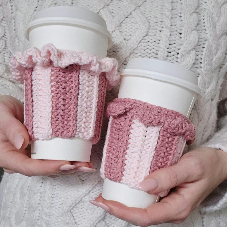 May include: Two white disposable coffee cups, each featuring a handmade crochet sleeve. The sleeves display vertical stripes in alternating shades of light pink and dusty rose, finished with a decorative ruffled edge at the top rim.