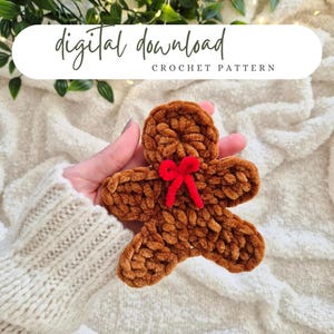May include: A brown crocheted gingerbread man with a red bow. The image includes the text "digital download" and "CROCHET PATTERN" in a white oval. The gingerbread man is held in a person's hand.
