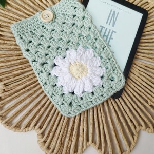 May include: A crocheted book cover in a light green color with a white daisy flower in the center. The cover is lying on a woven straw mat with a Kindle e-reader on top of it. The e-reader is displaying the text "Lakes Hockey Three In The"