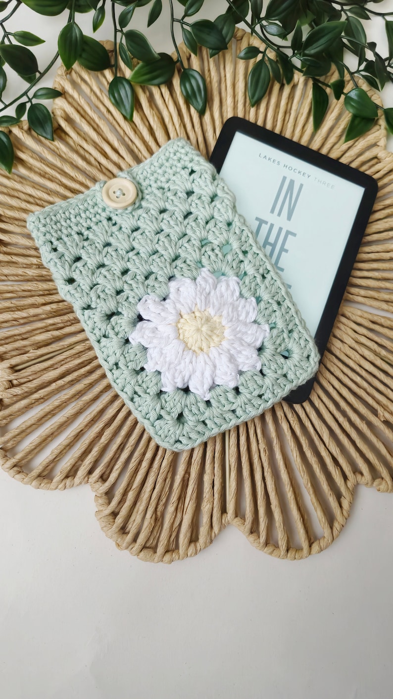 May include: A crocheted book cover in a light green color with a white daisy flower in the center. The cover is lying on a woven straw mat with a Kindle e-reader on top of it. The e-reader is displaying the text "Lakes Hockey Three In The"