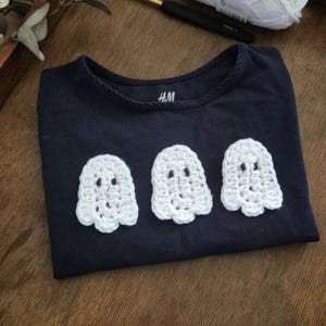 May include: Navy blue t-shirt with three white crochet ghost appliqu&eacute;s. The shirt has a round neckline and short sleeves. The brand label "H&M" is visible on the shirt. The shirt is folded on a wooden surface.