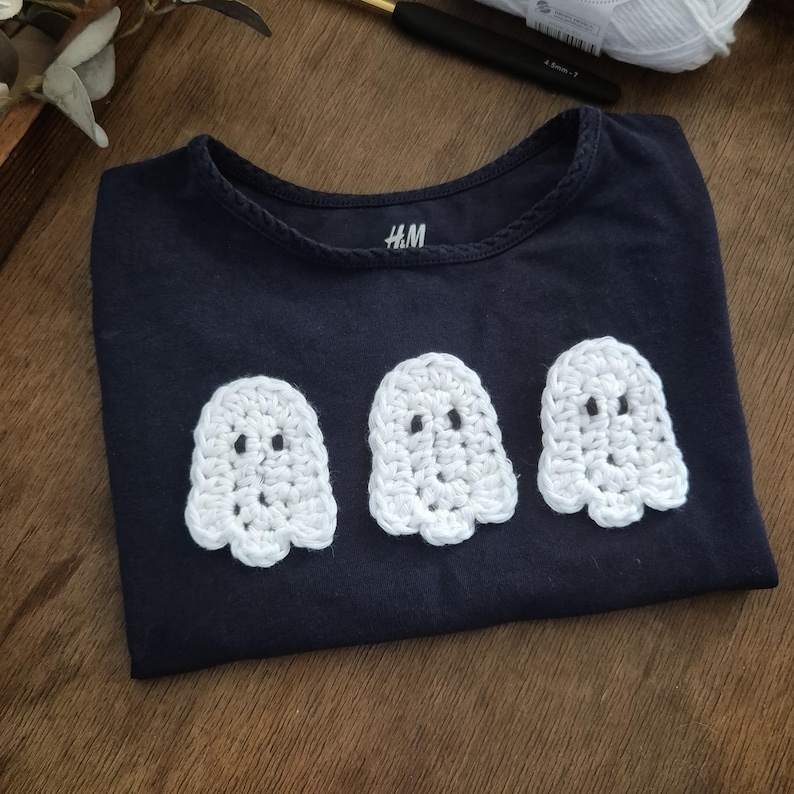 May include: Navy blue t-shirt with three white crochet ghost appliqu&eacute;s. The shirt has a round neckline and short sleeves. The brand label "H&M" is visible on the shirt. The shirt is folded on a wooden surface.