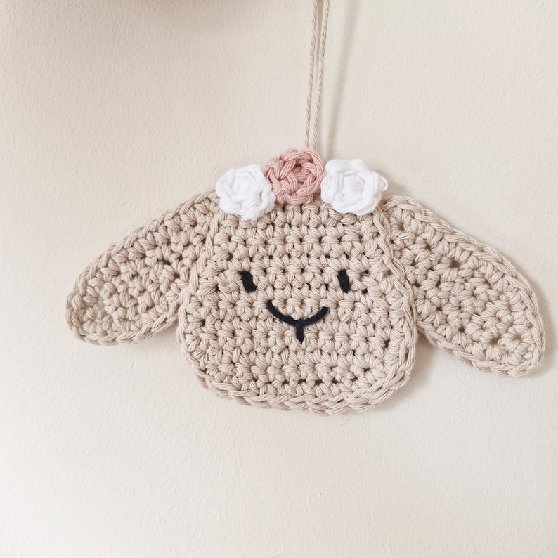 May include: A crocheted lamb ornament with a pink and white flower crown. The lamb is beige and has a black embroidered face.