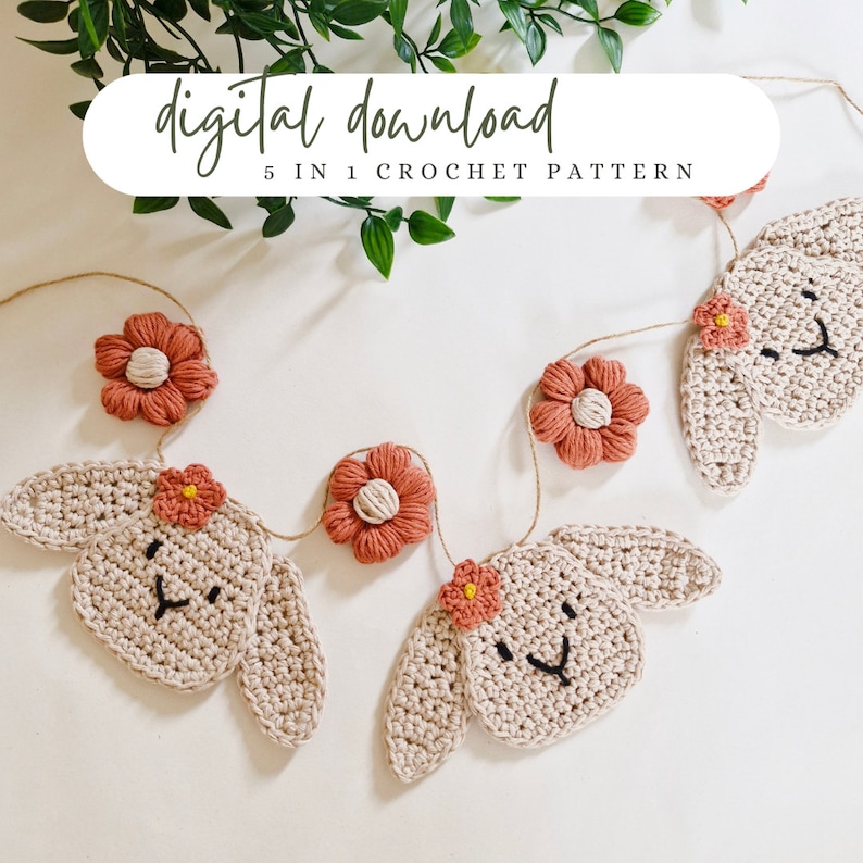 May include: A crochet garland with five bunny faces and five flowers. The bunnies are beige with black eyes and a black nose. The flowers are coral with a white center. The garland is strung with twine.