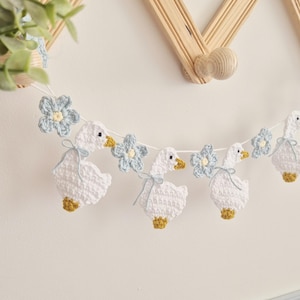 May include: A crochet garland featuring white geese with yellow beaks and feet, accented with light blue flowers. Each goose is tied with a thin light blue ribbon around its neck. The figures are connected by a white string, creating a decorative banner.