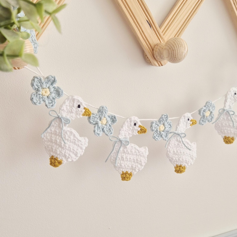 May include: A crochet garland featuring white geese with yellow beaks and feet, accented with light blue flowers. Each goose is tied with a thin light blue ribbon around its neck. The figures are connected by a white string, creating a decorative banner.