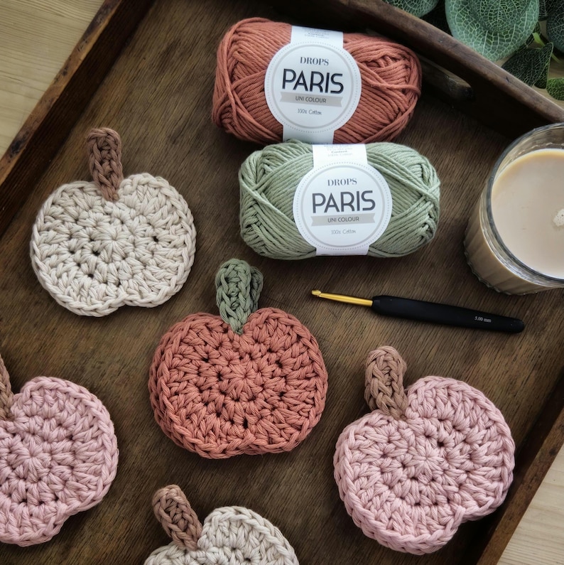May include: A wooden tray holds several crocheted pumpkin coasters in shades of pink, beige, and orange. Two skeins of yarn in matching colors and a crochet hook are also present. A lit candle sits in the background.
