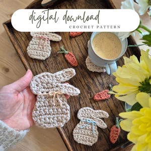 Può includere: Un vassoio di legno mostra disegni di coniglietti e carote all'uncinetto, insieme a una tazza di caffè. I coniglietti sono beige con fiocchi rosa o blu. Le carote sono arancioni con cime verdi. Il testo "digital download CROCHET PATTERN" è in alto.
