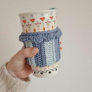 May include: A ceramic travel mug with a light blue crochet sleeve featuring a textured ribbed pattern and a scalloped top edge. The mug has a white base decorated with a repeating pattern of red and orange flowers, light blue dots, and black circles.