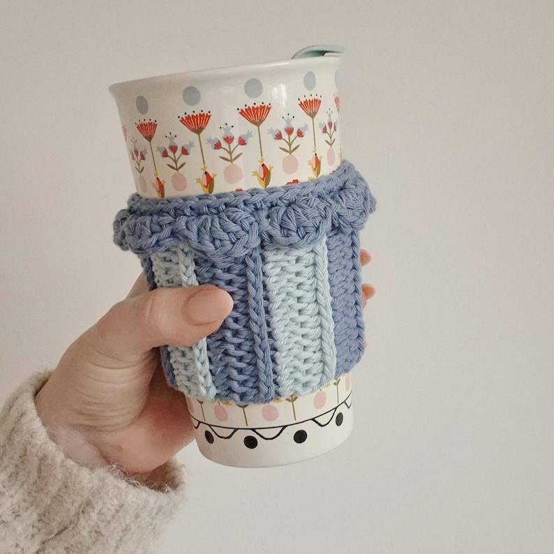 May include: A ceramic travel mug with a light blue crochet sleeve featuring a textured ribbed pattern and a scalloped top edge. The mug has a white base decorated with a repeating pattern of red and orange flowers, light blue dots, and black circles.