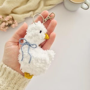 May include: A white fluffy pom-pom keychain shaped like a duckling, featuring a small yellow beak, yellow feet, and a light blue yarn bow tied around its neck. The keychain includes a silver-toned metal lobster clasp and a circular ring.
