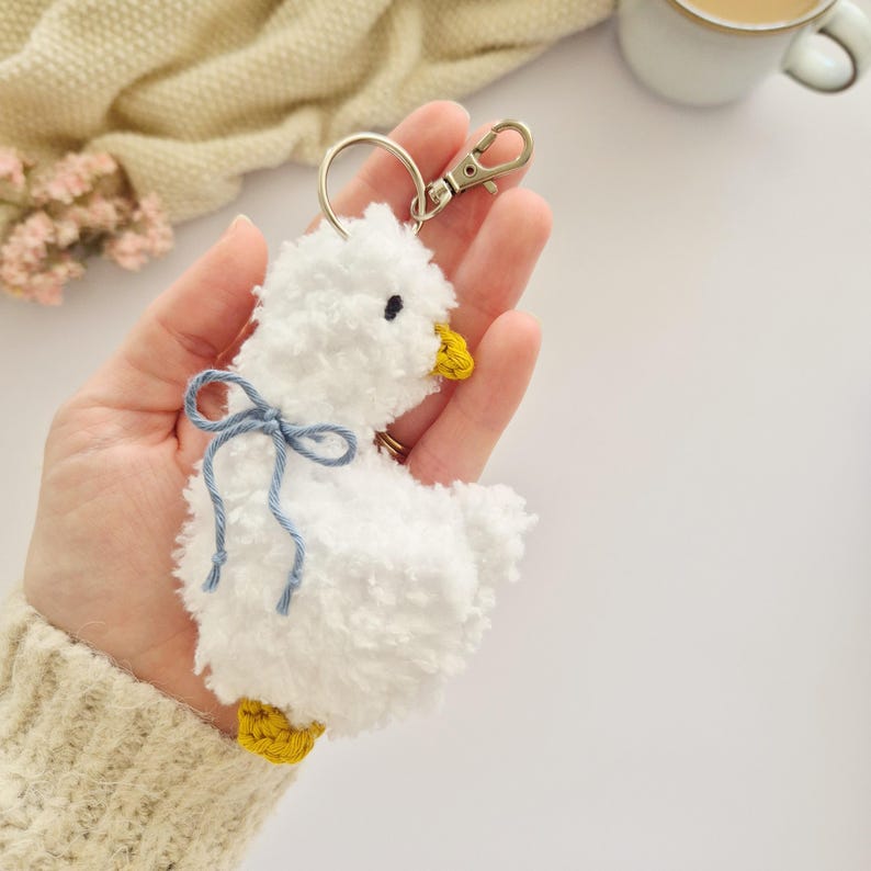 May include: A white fluffy pom-pom keychain shaped like a duckling, featuring a small yellow beak, yellow feet, and a light blue yarn bow tied around its neck. The keychain includes a silver-toned metal lobster clasp and a circular ring.