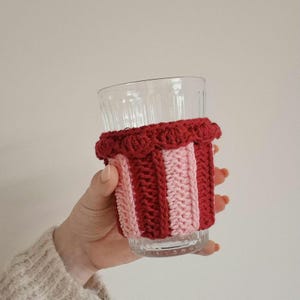 May include: A clear glass tumbler featuring a vertical striped crochet sleeve in alternating dark red and light pink yarn. The sleeve has a textured, ribbed knit pattern and a decorative scalloped edge along the top rim of the glass.