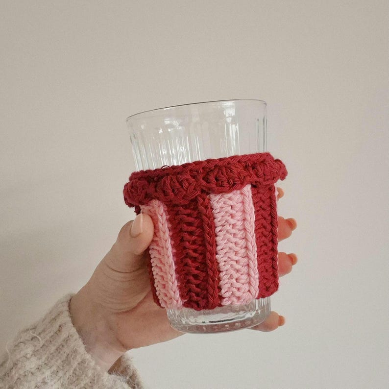 May include: A clear glass tumbler featuring a vertical striped crochet sleeve in alternating dark red and light pink yarn. The sleeve has a textured, ribbed knit pattern and a decorative scalloped edge along the top rim of the glass.