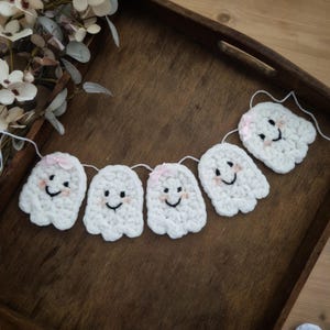 May include: A white crochet ghost garland with smiling faces and pink bows. The garland is strung on white thread and is displayed on a wooden tray. The ghosts are a cute and festive decoration.