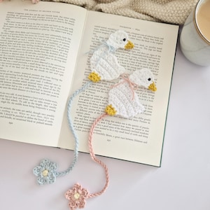 May include: Two crocheted white duck bookmarks with yellow beaks and feet. One duck has a light blue ribbon and a matching light blue crocheted flower at the end of its tail, while the other has a pink ribbon and a matching pink crocheted flower.