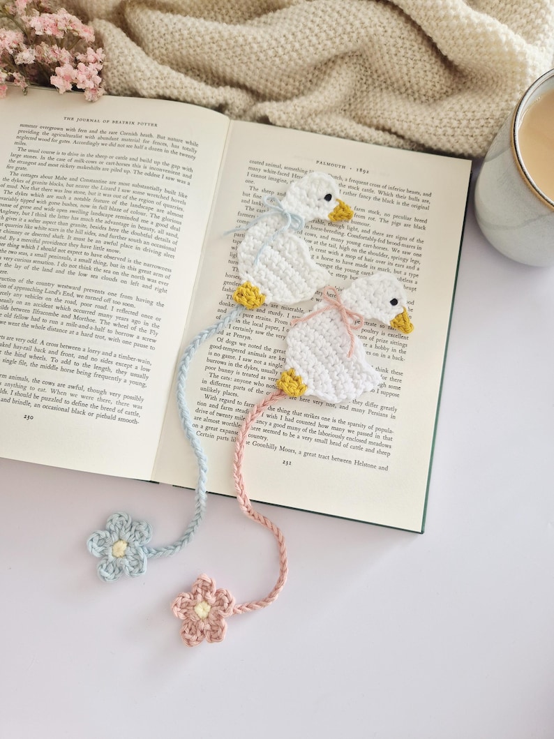 May include: Two crocheted white duck bookmarks with yellow beaks and feet. One duck has a light blue ribbon and a matching light blue crocheted flower at the end of its tail, while the other has a pink ribbon and a matching pink crocheted flower.