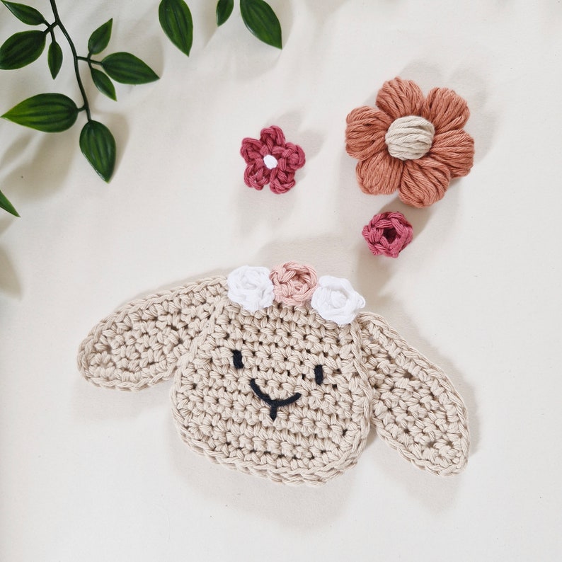 May include: A crocheted lamb with a brown face and white flowers on its head. The lamb is surrounded by pink and orange crocheted flowers.