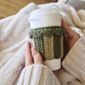 May include: A white disposable coffee cup featuring a removable crochet sleeve. The sleeve is crafted with alternating vertical stripes of sage green and cream yarn, finished with a decorative scalloped edge along the top rim of the cup.