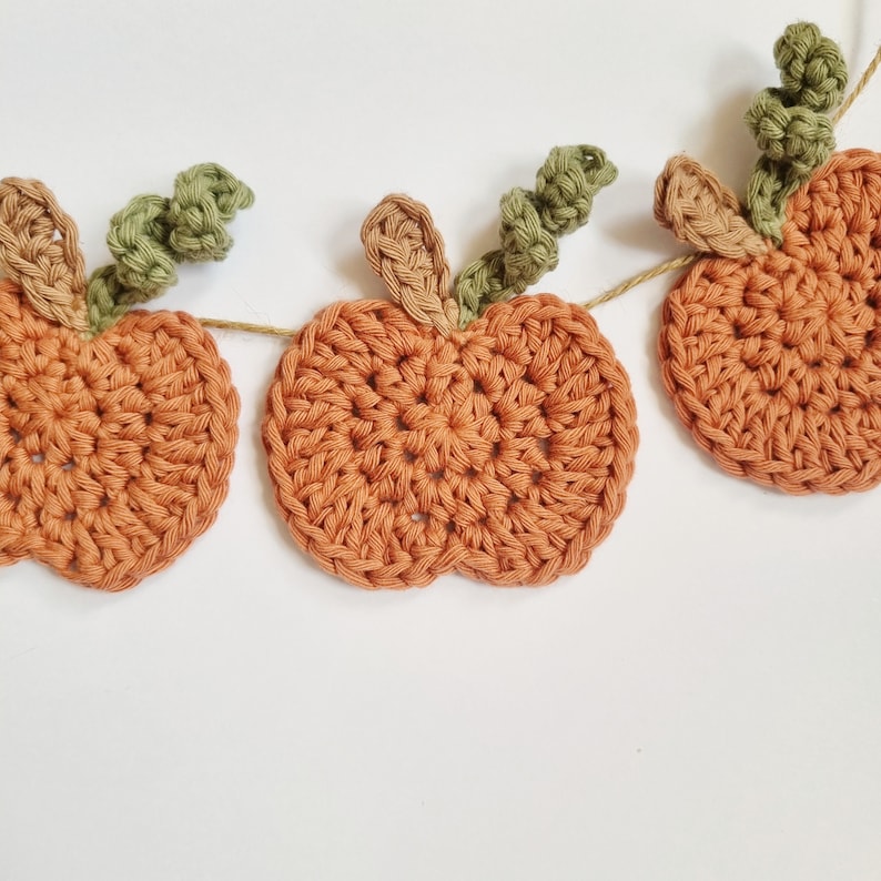May include: A garland of crocheted pumpkins with green leaves. The pumpkins are orange and the leaves are a light green.