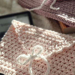 Puede incluir: Dos bolsos de ganchillo hechos a mano con forma de sobre, en rosa claro y malva. Cada bolso tiene un cierre anudado blanco. La puntada texturizada y los detalles de lazo sugieren un proyecto artesanal, ideal para peque&ntilde;os regalos o almacenamiento.
