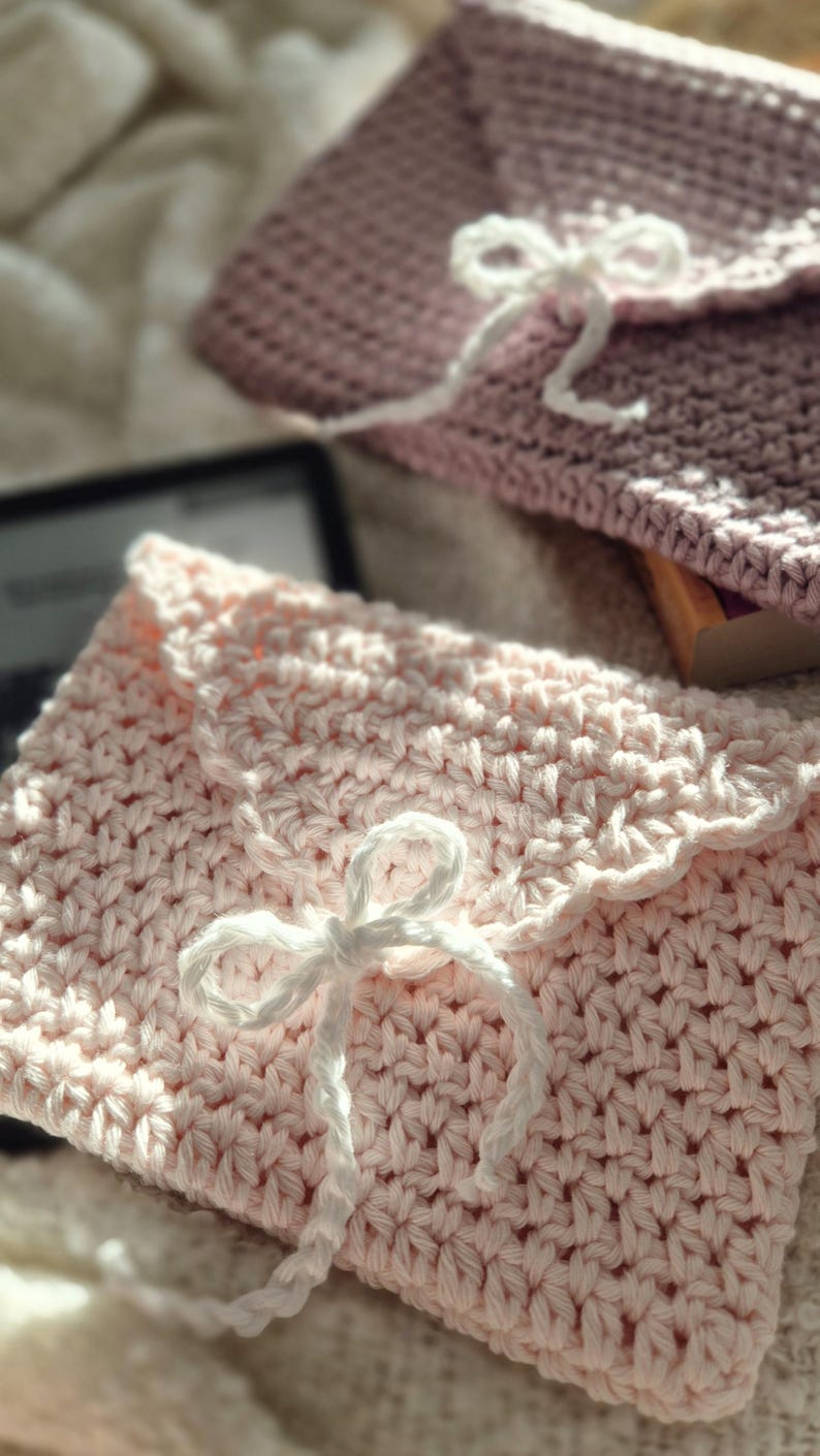 Puede incluir: Dos bolsos de ganchillo hechos a mano con forma de sobre, en rosa claro y malva. Cada bolso tiene un cierre anudado blanco. La puntada texturizada y los detalles de lazo sugieren un proyecto artesanal, ideal para peque&ntilde;os regalos o almacenamiento.