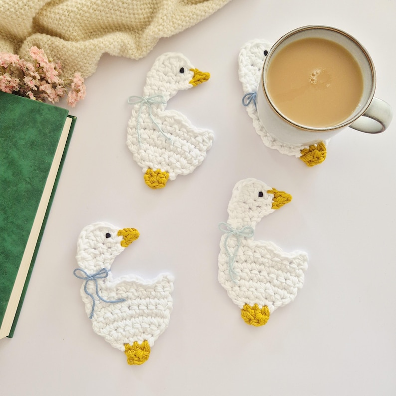 May include: Three white crochet goose coasters with yellow beaks and feet, each featuring a small light blue bow tied around the neck. One coaster is positioned under a ceramic mug filled with a light brown beverage, while two others lie flat.