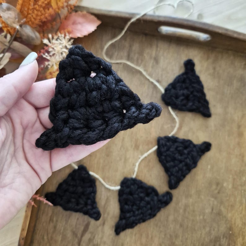 May include: A black crochet garland featuring witch hat shapes strung on a natural twine. The handmade garland is displayed on a wooden tray, perfect for Halloween or fall decor.