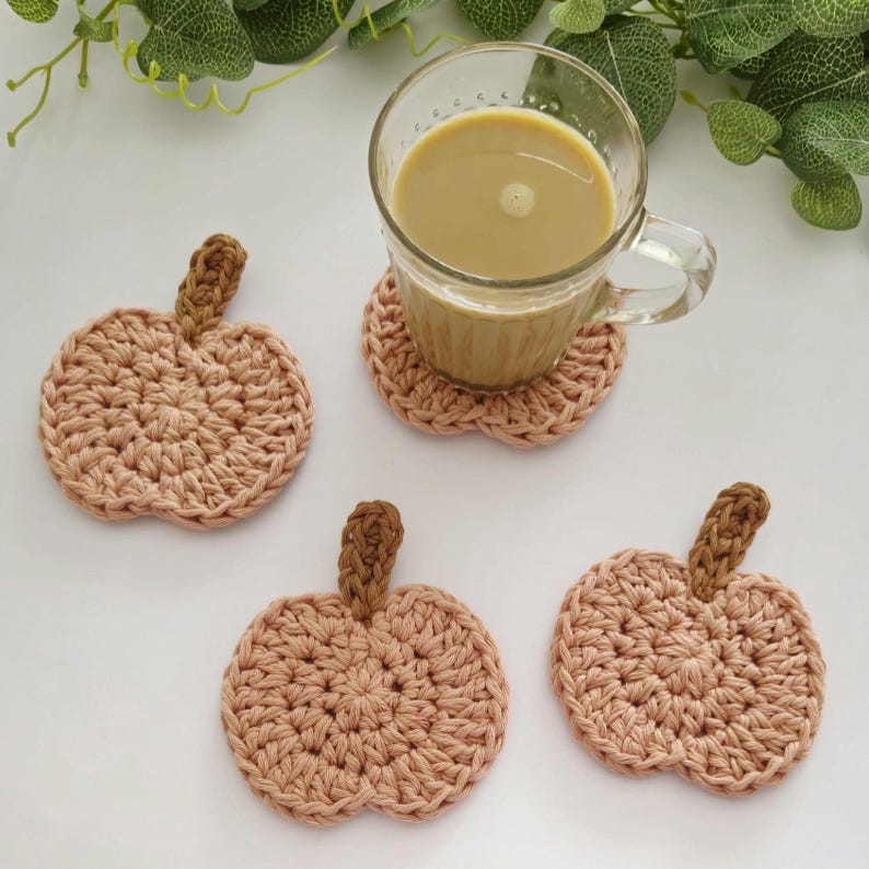 May include: Four handmade crochet pumpkin coasters in a light peach color with brown stems. One coaster is under a glass mug filled with a light brown liquid. The coasters are on a white surface.