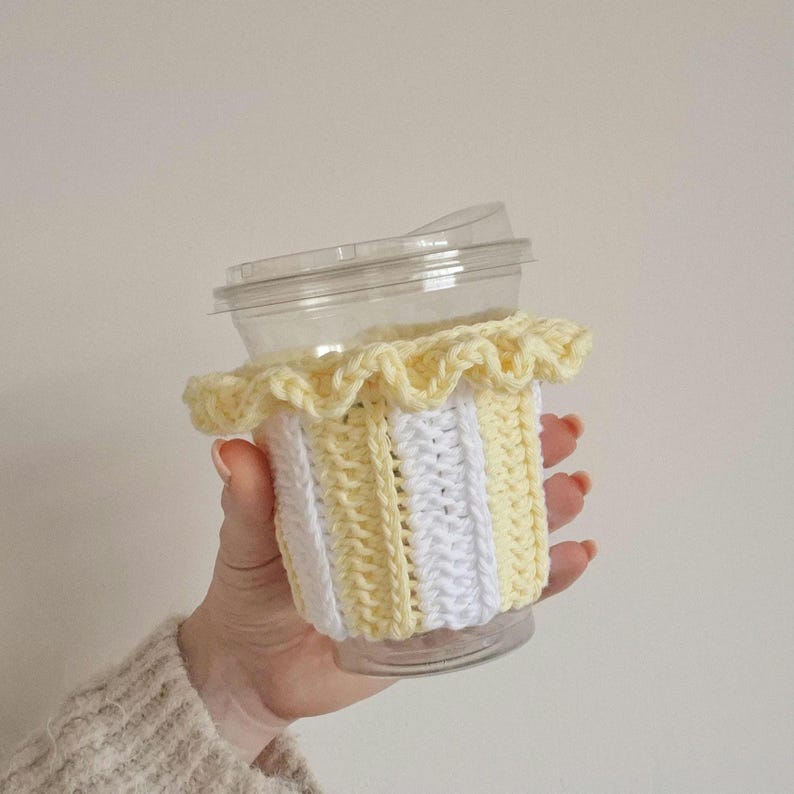 May include: A clear plastic cup featuring a removable crochet sleeve with vertical stripes in pale yellow and white. The sleeve includes a decorative ruffled edge at the top, providing a textured layer around the exterior of the beverage container.