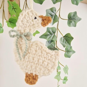 May include: A cream-colored crocheted goose ornament featuring a brown beak and feet, with a sage green yarn bow tied around its neck. The goose is crafted from thick yarn and includes a small loop at the top for hanging, showcasing a textured stitch.