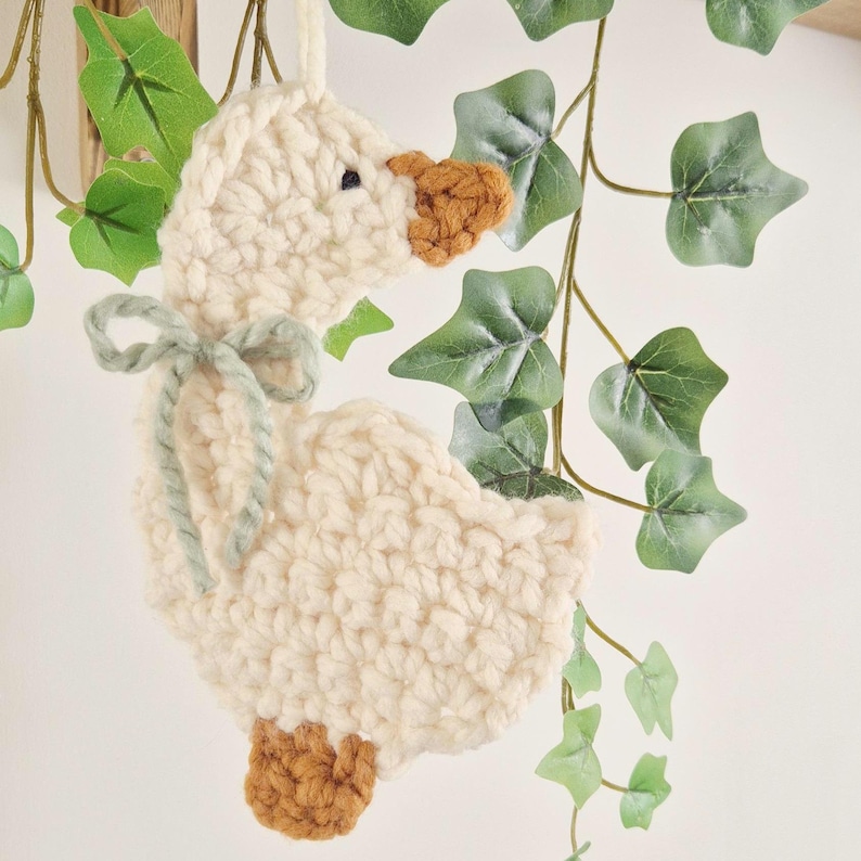 May include: A cream-colored crocheted goose ornament featuring a brown beak and feet, with a sage green yarn bow tied around its neck. The goose is crafted from thick yarn and includes a small loop at the top for hanging, showcasing a textured stitch.