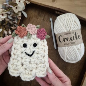 May include: A handmade, off-white crochet ghost with a smiling face and a floral crown. The ghost is held in front of a ball of cream-colored yarn with a label that reads "Yarnsmiths Create Super Chunky". A crochet hook is also visible.